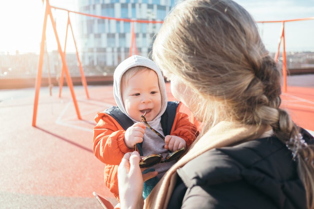 Mãe segura seu filho pequeno, ao colo, em uma pracinha ensolarada, num dia frio de inverno.